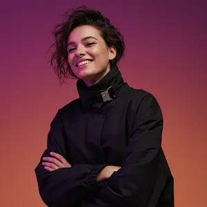 A smiling woman with short, wavy hair and crossed arms, wearing a black high-collared jacket against a purple-to-orange gradient background.