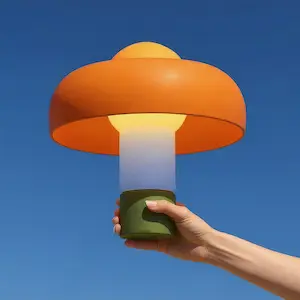 A hand holding up a portable, mushroom-shaped lamp with a bright orange shade and green base against a clear blue sky. 